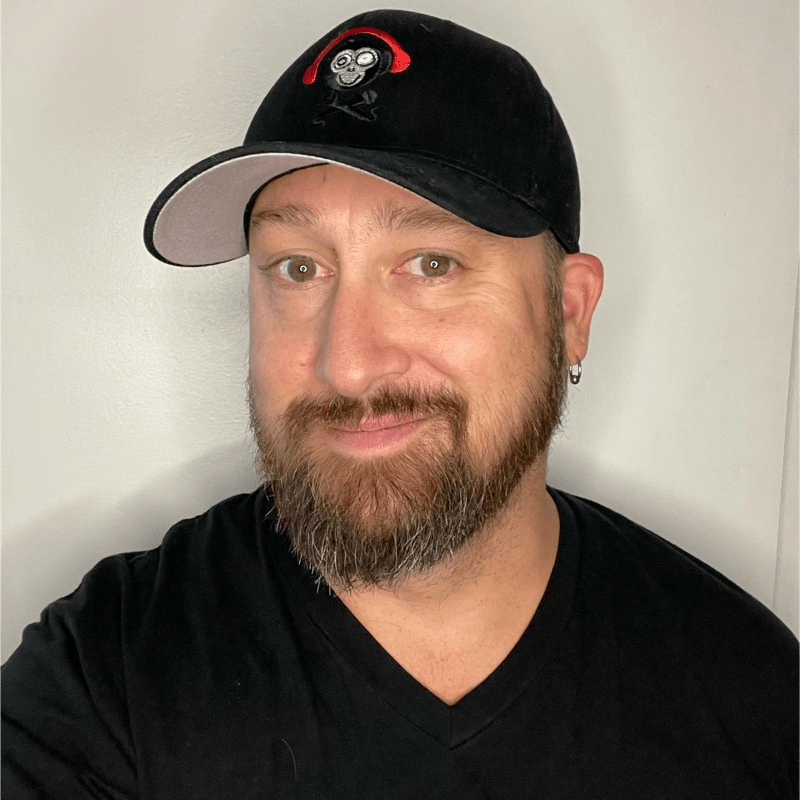 A bearded man wearing a black T-shirt and a black cap with a monkey logo, smiling slightly in front of a plain light-colored wall, appears ready to host the Invigor Medical Podcast.