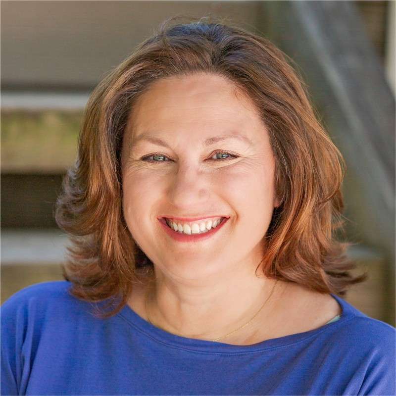 A woman with shoulder-length brown hair and a blue top smiles warmly at the camera, radiating the calm confidence of someone who practices Yoga for Longevity. Wooden steps and greenery are softly blurred in the background.
