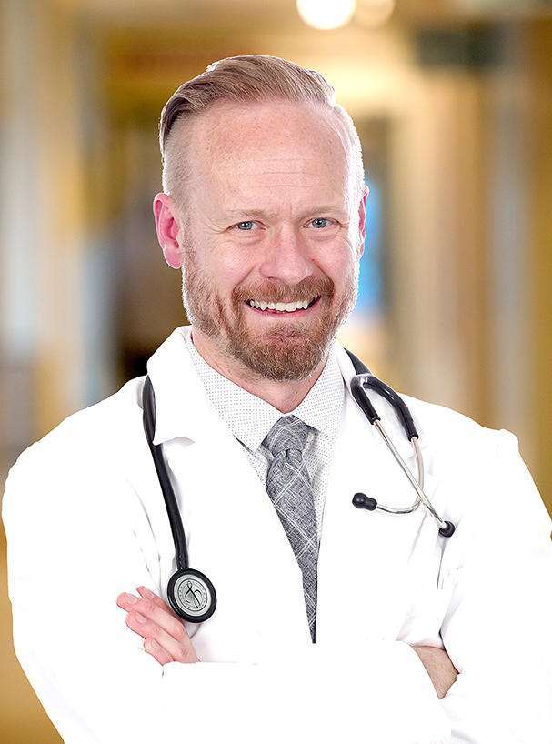 A smiling male doctor with a beard and mustache, wearing a white lab coat, tie, and stethoscope, stands with arms crossed in a brightly lit hallway—ready to discuss causes and treatments for ED.