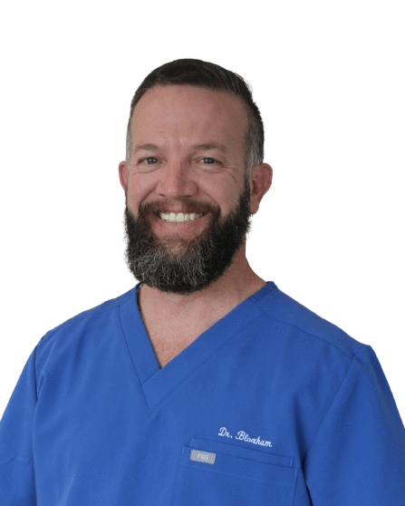 A smiling man with a beard wearing blue medical scrubs embroidered with “Dr. Bonham” stands against a plain background, ready to share expert tips on optimizing sleep for better overall health.
