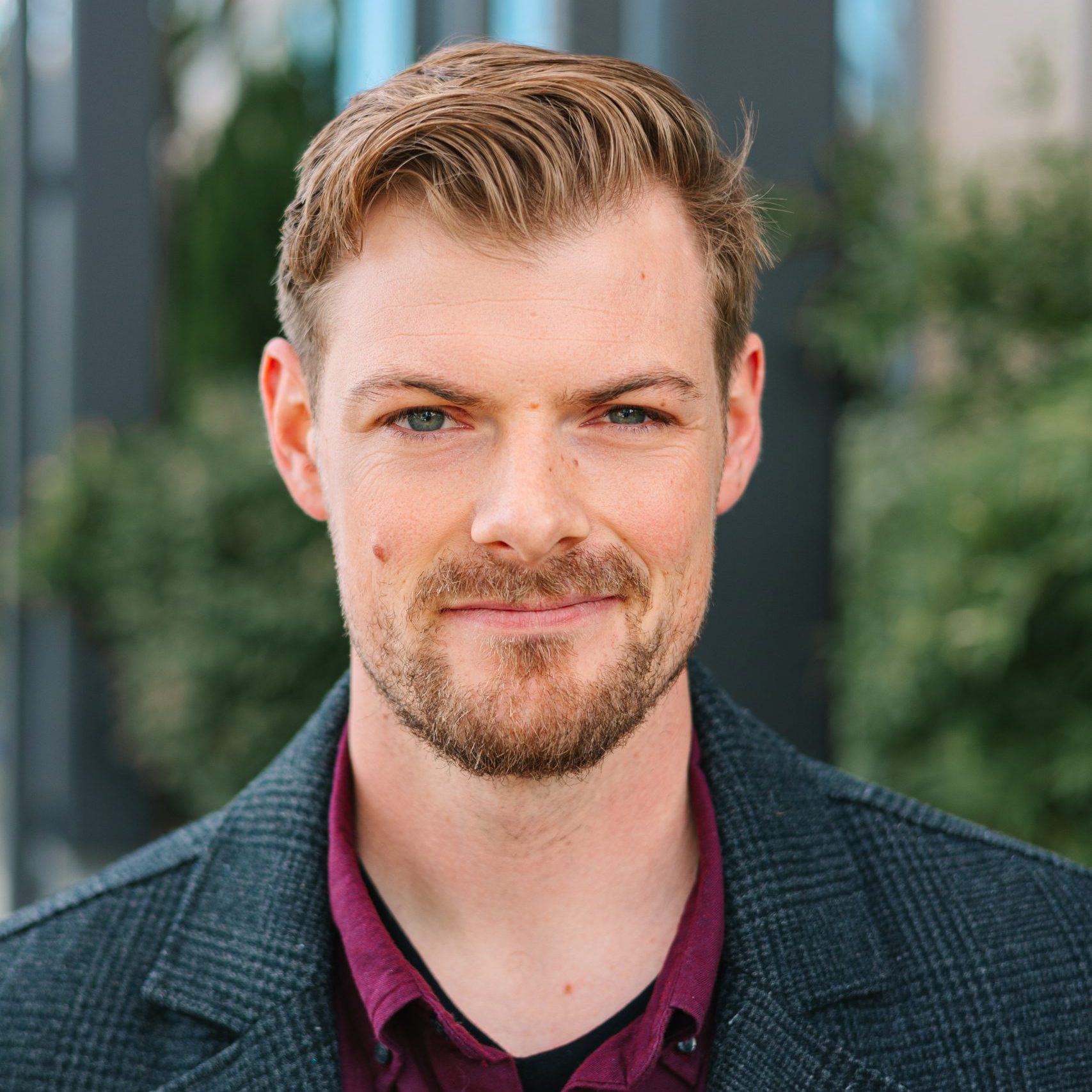 A man with short, light brown hair and a trimmed beard is smiling slightly. He is wearing a dark jacket over a burgundy shirt, standing outdoors with greenery in the background, looking confident after his recent weight loss.