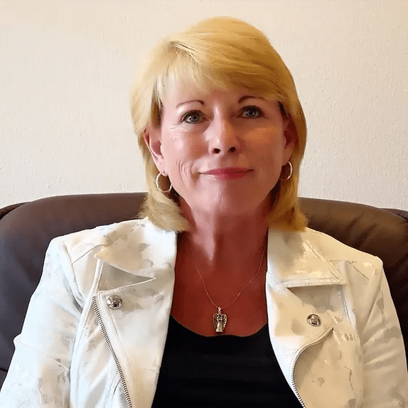 A woman with short blonde hair, wearing a white jacket over a black top and a silver necklace, sits on a dark brown couch, smiling softly at the camera—her calm demeanor reflecting the balance she’s found through bioidentical hormone replacement therapy.