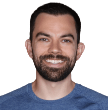 A man with short dark hair, a trimmed beard, and mustache, wearing a blue crew-neck shirt, smiles at the camera against a plain background, appearing ready for his blood flow restriction workout.