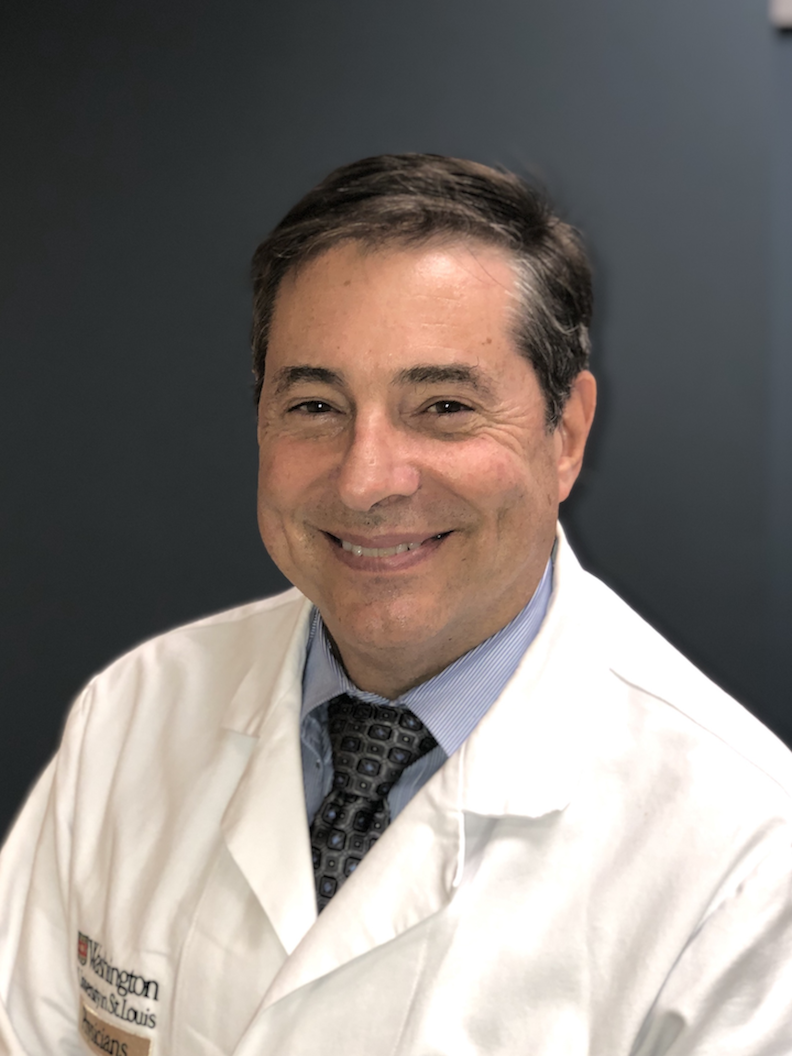 A man in a white doctor’s coat and patterned tie, specializing in brain health and Alzheimers care, smiles at the camera, seated against a plain dark background.