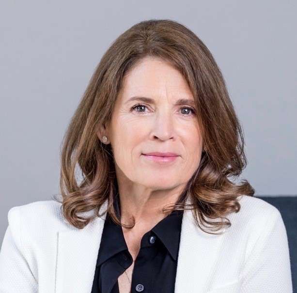 A woman with shoulder-length brown hair, wearing a white blazer over a black shirt, sits in front of a neutral gray background, looking directly at the camera with calm mental strength.