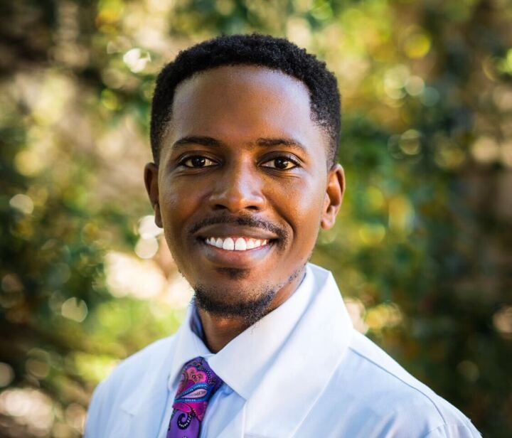 A smiling man wearing a white lab coat and a colorful tie stands outdoors in front of blurred green foliage, embodying the dedication of professionals advancing GLP-1 therapies.