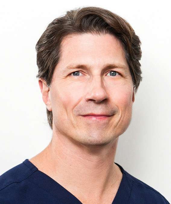 Dr. Jay Calvert, a man with light skin and brown hair wearing a dark blue top, smiles slightly as he poses in front of a plain white background.