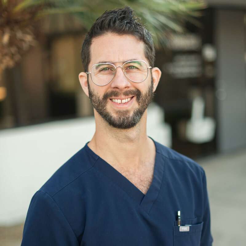 A man with short dark hair, a beard, and glasses smiles at the camera while wearing navy blue scrubs and standing outdoors. A pen is visible in his chest pocket, reflecting his commitment to healing through innovative therapies like PRP.