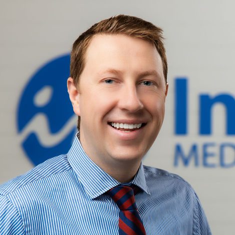 A smiling man with short light brown hair wearing a blue striped shirt and red-striped tie stands in front of a blurred background with blue and white text and logo, ready to give an introduction on the future of medicine and the role of peptides.