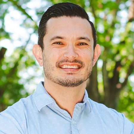 A man with short dark hair and a trimmed beard smiles outdoors, wearing a light blue collared shirt. Green trees and sunlight create a blurred natural background, adding warmth as he discusses common Bedroom Myths with confidence.