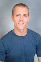 A man with short light brown hair, wearing a blue performance t-shirt, is smiling slightly and facing the camera against a plain grey background.