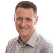 A man with short brown hair, wearing a light gray, striped button-up shirt, is smiling and facing the camera against a plain white background, exuding the invigorating energy of cold water immersion.