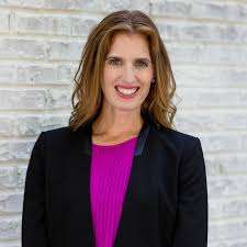A woman with wavy brown hair, wearing a black blazer over a bright pink top, stands smiling in front of a white brick wall, radiating confidence and positivity about gut and mental health.