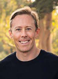 A smiling man with short blond hair wearing a black shirt stands outdoors in front of blurred green trees, radiating positivity on his journey of healing and recovery.