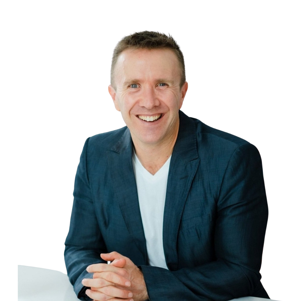 A smiling man with short hair wearing a navy blazer over a white shirt sits at a white table, hands clasped, ready to discuss his daily routine. The background is plain white.