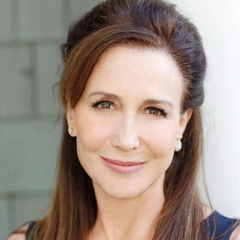 A woman with long brown hair, styled back, and wearing stud earrings, smiles softly at the camera. She is dressed in a dark top, and the softly blurred background gently highlights Emily Gold Mearss poised demeanor.