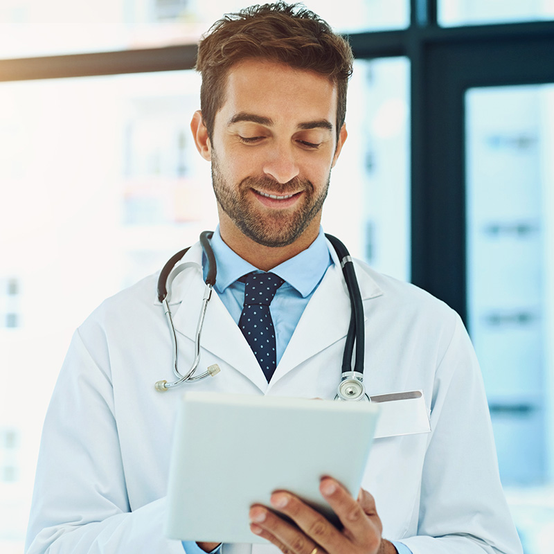 A male doctor with a beard, wearing a white coat and stethoscope, smiles while looking at a tablet device in a bright medical office.