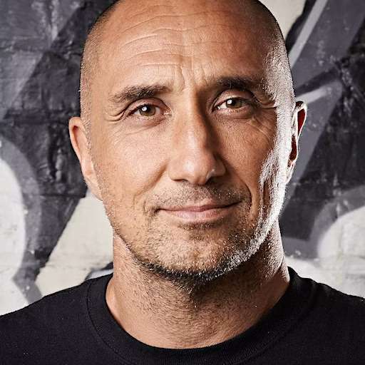 A bald man with a slight smile and stubble, identified as Vic Verdier, is wearing a black shirt and standing in front of a graffiti-covered wall. The image is a close-up portrait with soft lighting.