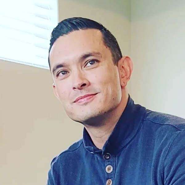 A man with short dark hair, wearing a dark blue button-up shirt, sits indoors with a slight smile, in front of a light-colored wall and a window with blinds, possibly discussing topics like erectile dysfunction.