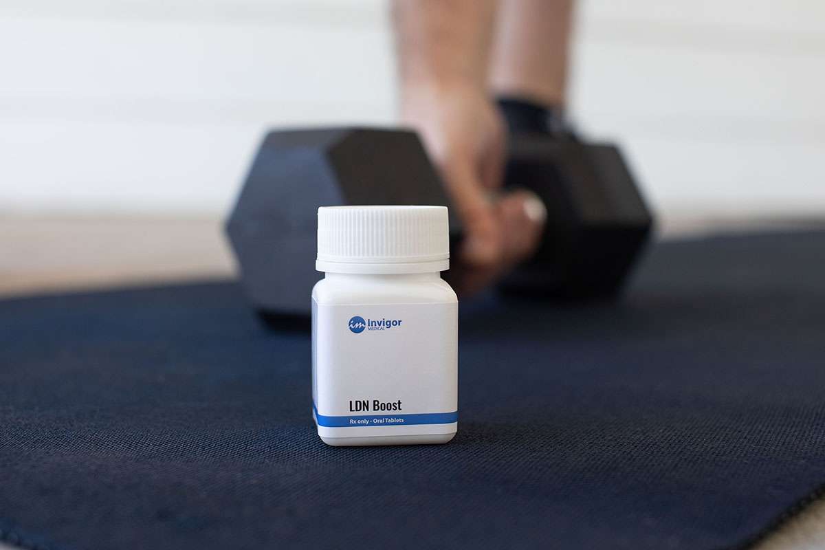 A white bottle labeled LDN Boost sits on a dark exercise mat, with a blurred background showing a hand gripping a black dumbbell—perfect for those looking to buy Low Dose Naltrexone and support their fitness goals.