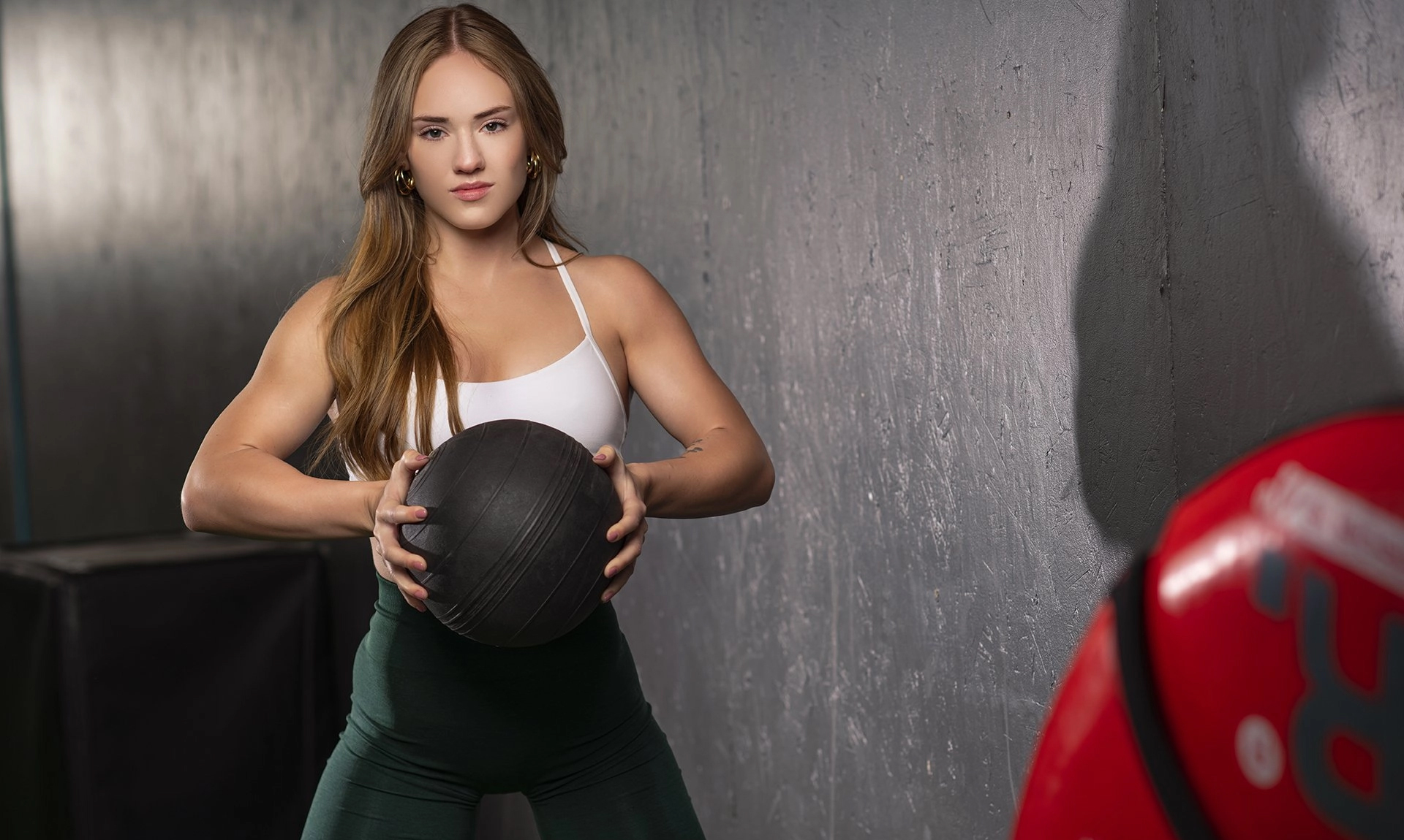 A young woman in a white tank top and green leggings demonstrates proper gym etiquette, holding a black medicine ball with both hands in a gym, standing near a gray wall with equipment in the background.