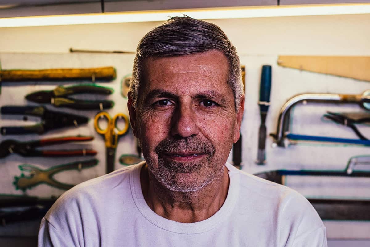 A middle-aged man with gray hair and a trimmed beard, an advocate for sexual health, wears a white shirt and smiles softly, standing in front of a wall with various hand tools hanging in a workshop.