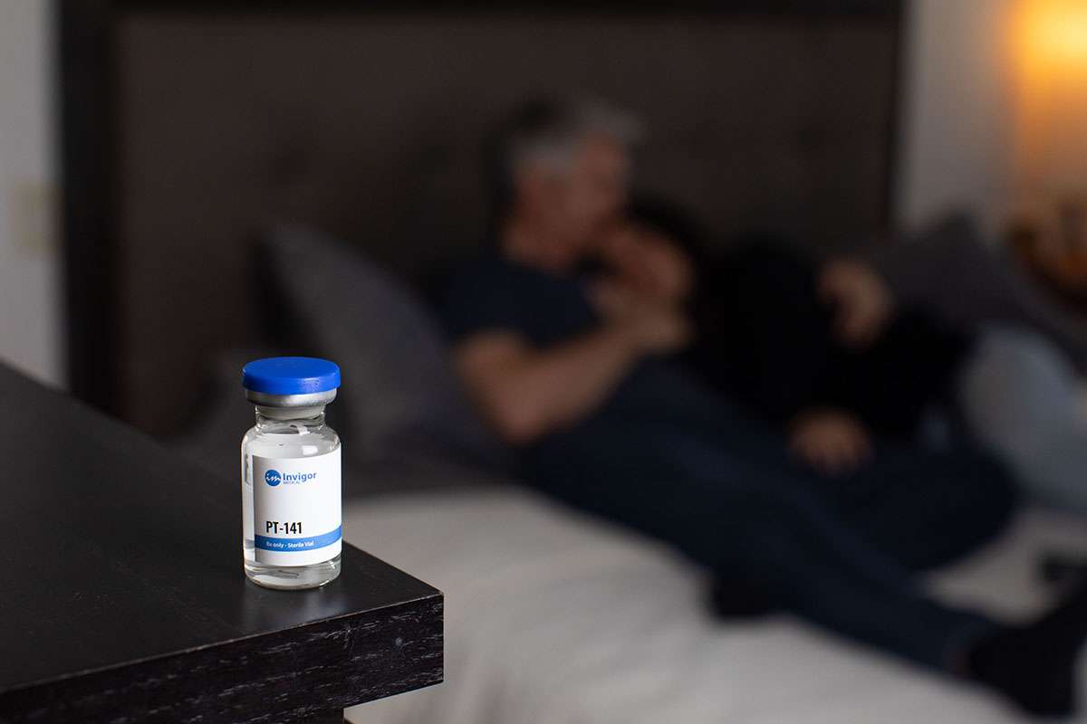 A vial labeled PT-141 sits on a bedside table, with a blurred couple cuddling on a bed in the background, suggesting romance—and inviting viewers to consider where to buy PT-141 injections for similar intimate moments.