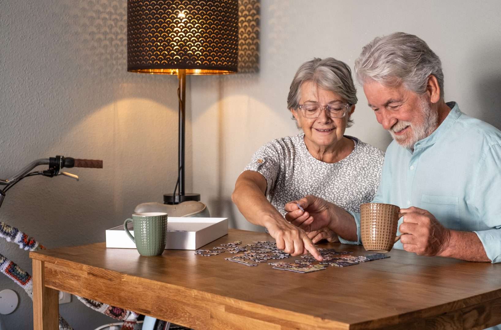 Two older adults sit at a wooden table, smiling and working on a jigsaw puzzle together to boost their mental acuity. Each holds a mug, with a lamp and bicycle in the background, creating a cozy atmosphere.