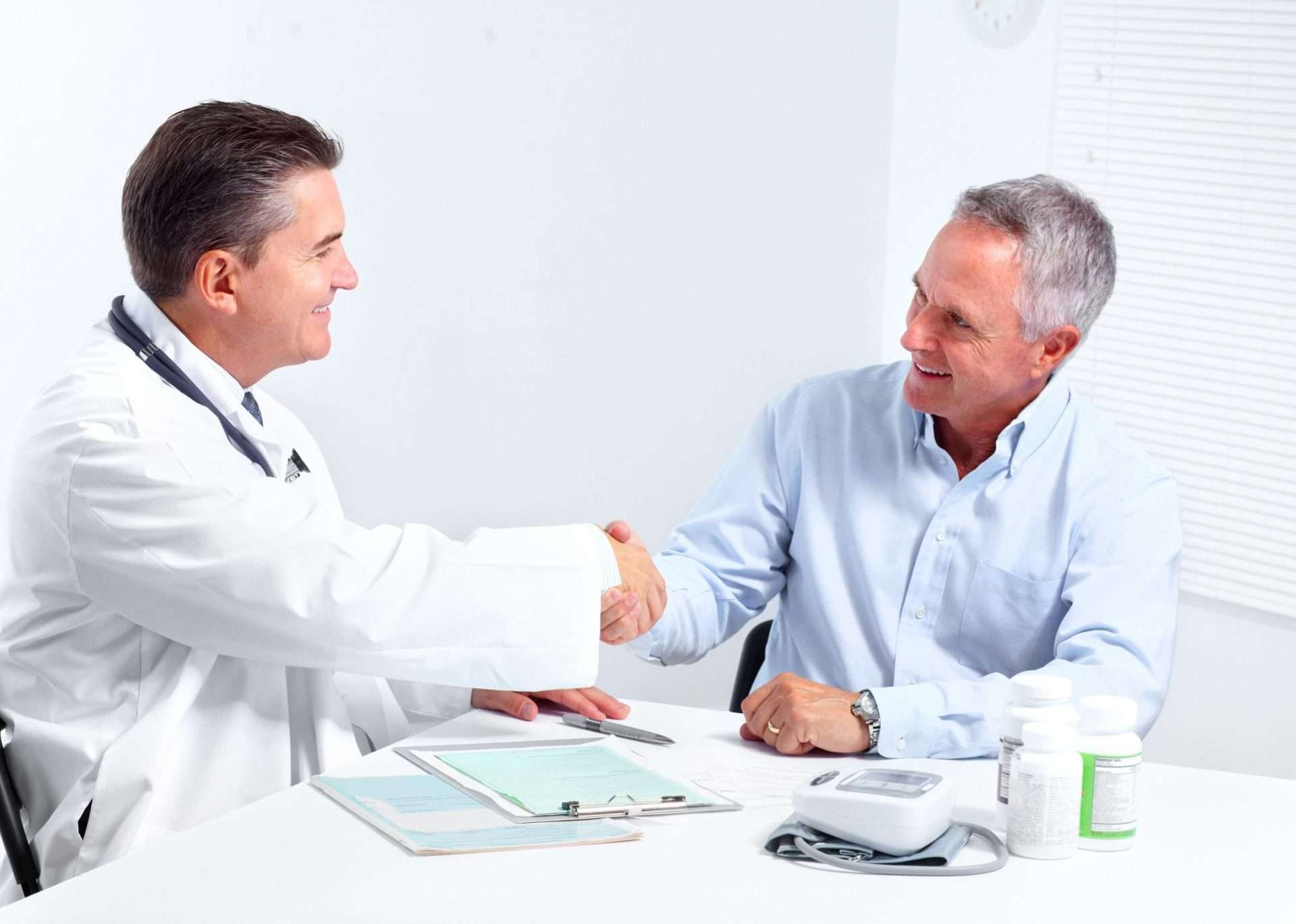 A doctor and an older male patient are sitting at a desk in one of the mens health clinics, smiling and shaking hands. Medical forms, a blood pressure monitor, and medication bottles are on the table in this bright, clinical setting.