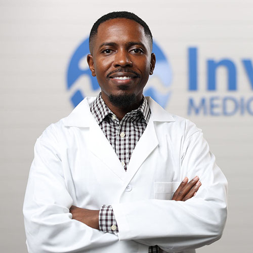 A man wearing a white lab coat and checkered shirt stands with arms crossed, smiling, in front of a blurred background with partial text and a blue logo.