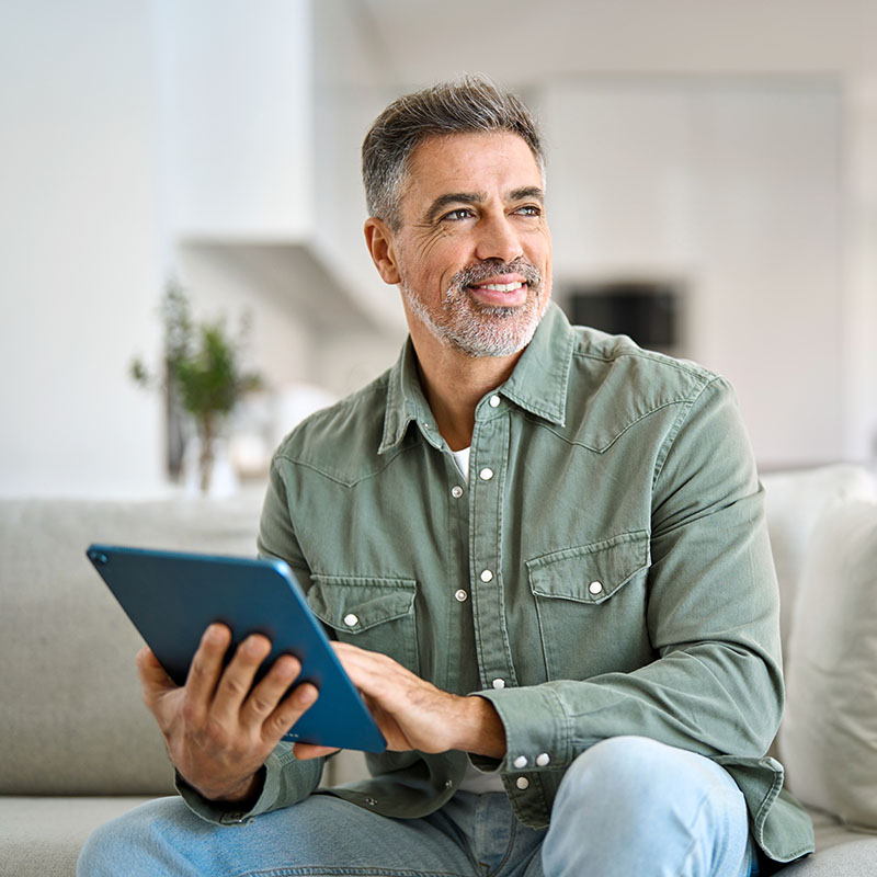 A middle-aged man with gray hair and a beard, wearing a green shirt, sits on a couch holding a tablet and smiling, in a bright and modern living room.