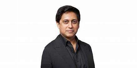 Dr. Srini Pillay, a man with short black hair, wearing a black suit and black shirt, stands against a plain white background, looking at the camera with a neutral expression.