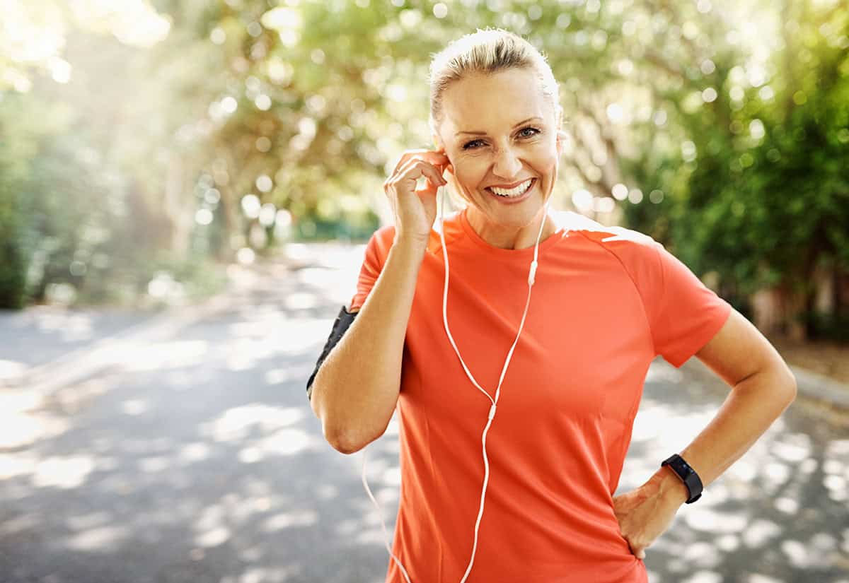 A smiling woman in an orange shirt stands outdoors on a sunny day, wearing earphones and a fitness tracker, with one hand on her hip and the other adjusting her earphones. Trees line the background.